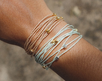 May include: A stack of colorful friendship bracelets with gold and silver beads. The bracelets are made of thin, braided cords.