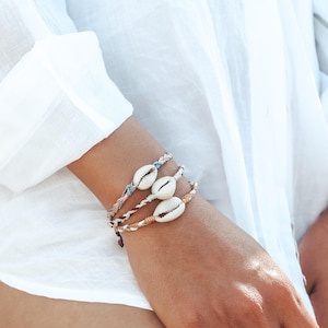 Può includere: Tre braccialetti intrecciati con conchiglie di ciprea. I braccialetti sono nei toni del marrone, rosa e blu. Le conchiglie sono bianche. I braccialetti sono indossati su un polso, con una camicia bianca sullo sfondo.