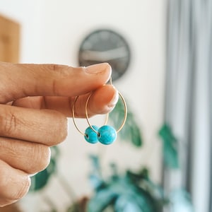 May include: Gold hoop earrings with turquoise blue beads. The earrings are held up by a hand, showcasing the jewelry against a blurred background of a room with plants and a clock.