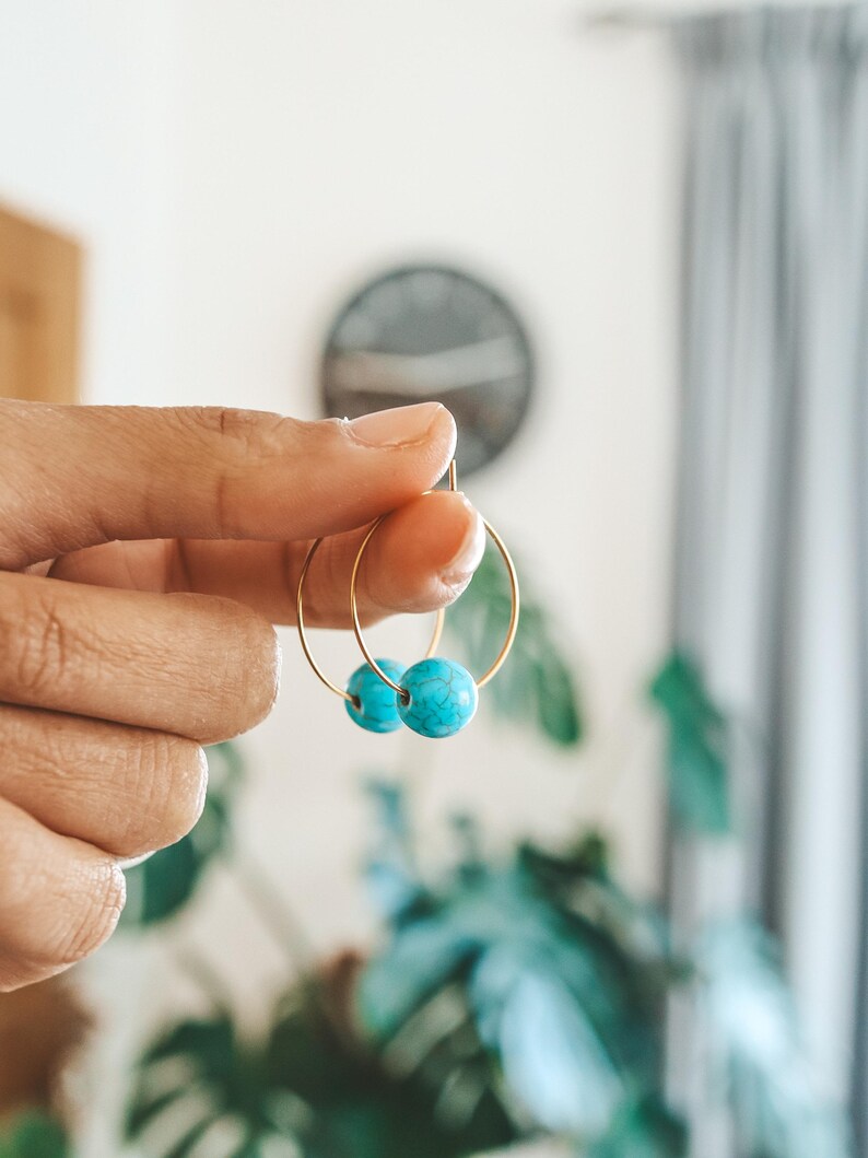 May include: Gold hoop earrings with turquoise blue beads. The earrings are held up by a hand, showcasing the jewelry against a blurred background of a room with plants and a clock.
