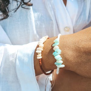 May include: Two handmade bracelets. One bracelet features off-white shell-like beads on a brown cord. The other bracelet has light blue and white shell-like beads on a white cord. Both bracelets are worn on a person's wrist.