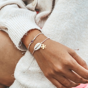 May include: Two bracelets on a wrist. The top bracelet is a thin white cord with a small pink stone charm. The bottom bracelet is a thicker white cord with a white seashell charm and a gold four-leaf clover charm.