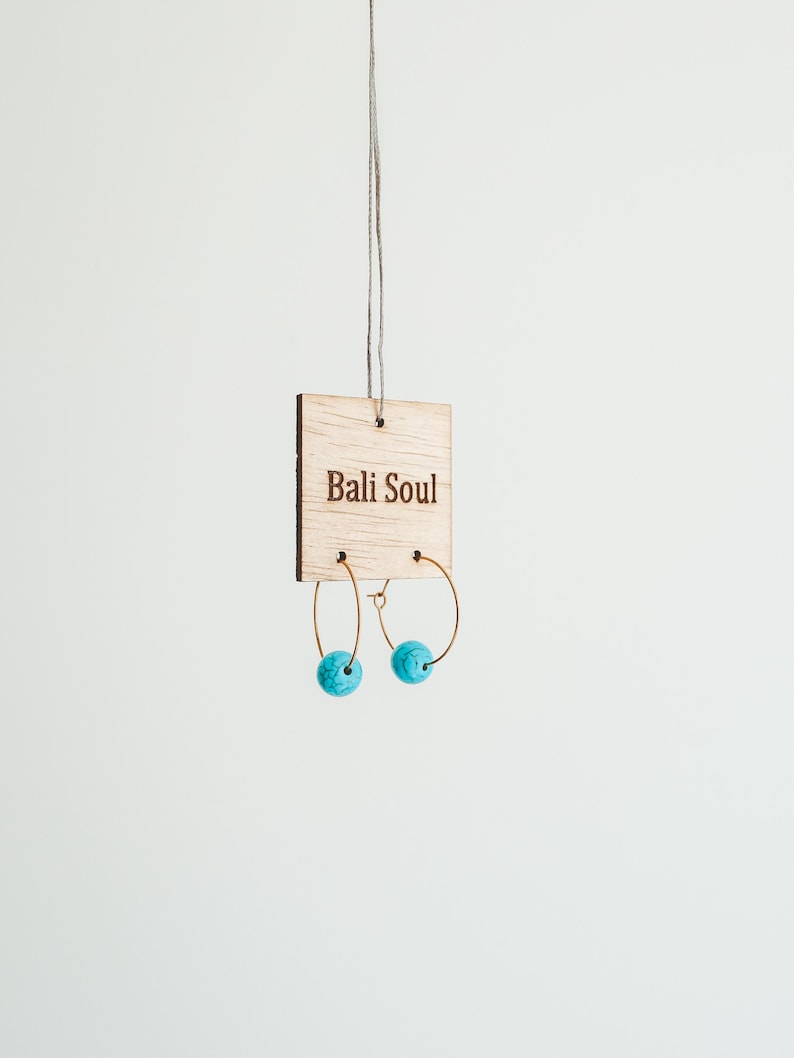 May include: A pair of gold-colored hoop earrings with turquoise blue spherical beads. The earrings are displayed on a wooden tag that reads "Bali Soul". The earrings hang from a thin string against a white background.