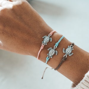 Könnte beinhalten: Drei Freundschaftsarmbänder mit silbernen Schildkrötenanhängern. Die Armbänder sind rosa, blau und braun.