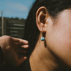 May include: A close-up shot of a gold hoop earring with a small, round, turquoise bead. The earring is worn on a person's ear. The background is blurred, with a hint of blue sky and greenery.