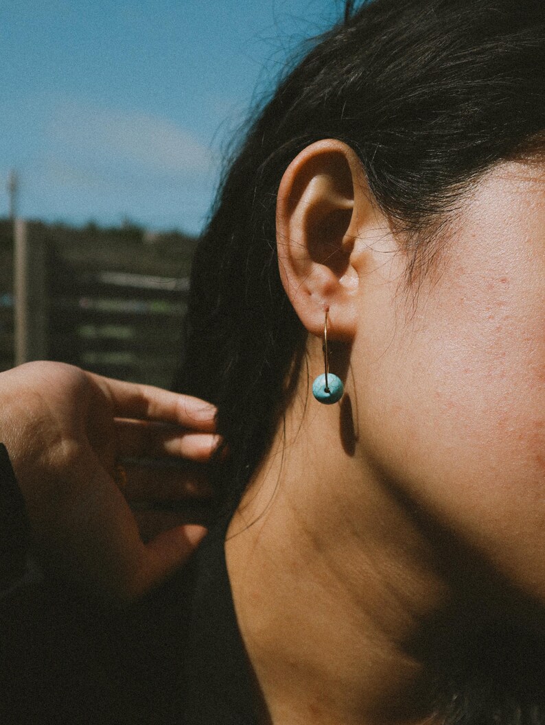 May include: A close-up shot of a gold hoop earring with a small, round, turquoise bead. The earring is worn on a person's ear. The background is blurred, with a hint of blue sky and greenery.