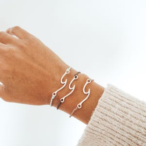 May include: Three bracelets with a silver wave charm on a person's wrist. The bracelets are made of brown, light blue, and white cord.