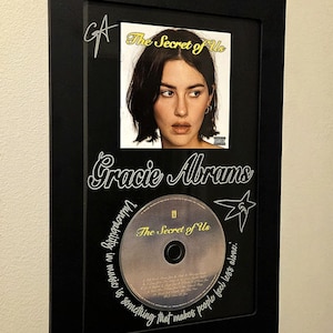 May include: Framed CD and album art for Gracie Abrams' "The Secret of Us." The black frame holds a photo of the artist and the CD. The album title and artist's name are displayed in white script. A star graphic is also included.