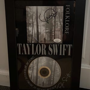 May include: A framed black and white photo of a forest with a CD in the bottom right corner. The CD has a forest image and the text "THERE IS NO ONE BETTER / TAYLOR SWIFT /  REYNOLDS 80 CSTL." The frame has the text "TAYLOR SWIFT" and "FOLKLORE" in white lettering.