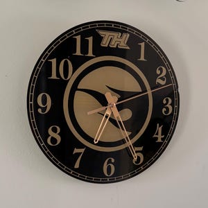 May include: A black and gold clock with a gold eagle logo and the letters "TH" in the top center. The clock face has gold numbers 1 through 12 and gold hands.