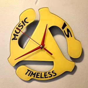 May include: A yellow wall clock shaped like a 45 RPM record adapter. The clock has red hands and the words "MUSIC IS TIMELESS" in black lettering. The clock is mounted on a light gray wall.