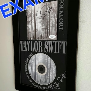 May include: A black framed display featuring a Taylor Swift Folklore CD and album art. The piece includes a signature, the text FOLKLORE, TAYLOR SWIFT, and the quote JUST BE YOURSELF, THERE IS NO ONE BETTER, along with a small authentication sticker.