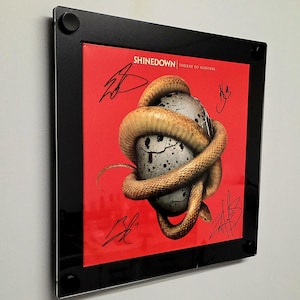 May include: Framed album art for Shinedown's "Threat to Survival." The artwork features a snake coiled around a cracked egg on a red background. The frame is black, and the album cover is signed.
