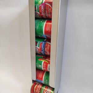 May include: A silver metal can dispenser holding several cans of tomato products. The cans are red and green with Italian text. The dispenser is designed to hold the cans in a vertical row.