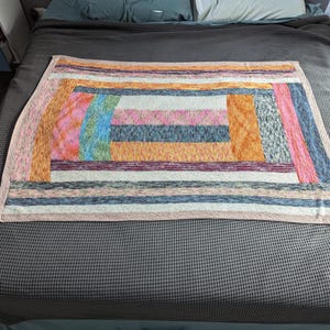 Op de afbeelding: Een veelkleurige deken met een patchwork-ontwerp. De deken heeft roze, oranje, blauwe en grijze tinten. De deken is gemaakt van een zacht materiaal, waarschijnlijk voor warmte en comfort. Afmetingen van de deken zijn niet zichtbaar.