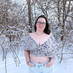 May include: A gray wrap top with a purple floral pattern. The top is worn with light blue jeans. The background features snow-covered trees and a snowy ground.