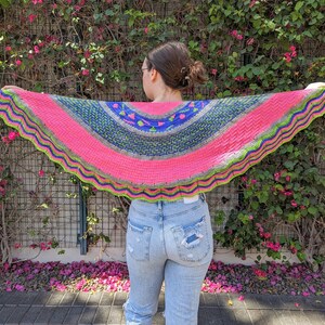 Könnte beinhalten: Ein rosa, blauer, grüner und grauer Häkel-Schal mit einem gewellten Rand. Der Schal ist über die Schultern einer Person gelegt, die vor einer Wand aus grünen Ranken mit rosa Blüten steht.