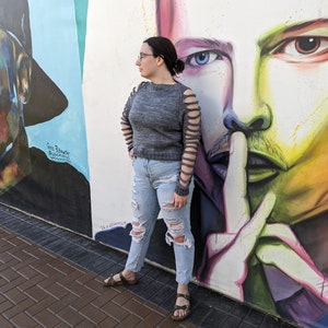 May include: A woman wearing a gray sweater with cut-out sleeves and distressed blue jeans stands in front of a wall with colorful murals. The murals feature two faces, one with a blue eye and the other with a brown eye.