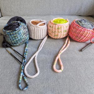 Könnte beinhalten: Vier handgefertigte Garnkörbe in Blau, Beige, Rosa und Rot, jeder mit Garn gefüllt. Stricknadeln und ein Häkelhaken sind in der Nähe der Körbe platziert. Die Körbe haben Griffe und sind aus Strickmaterial. Die Garnfarben in den Körben variieren.