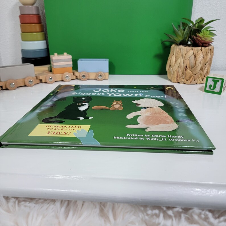 Pu&ograve; includere: Un libro verde con il titolo "Jake e lo sbadiglio pi&ugrave; grande di sempre!" La copertina presenta un gatto nero, uno scoiattolo marrone e un golden retriever. Il libro ha un adesivo che dice "Garantito per farti sbadigliare!"
