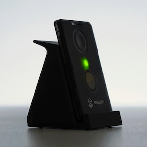 Op de afbeelding: Een zwarte smartphone met een groen licht, rustend op een zwarte driehoekige standaard. De telefoon heeft twee cirkelvormige elementen en een logo. De standaard is schuin geplaatst en ondersteunt het apparaat. De achtergrond is een verloop van wit en grijs.