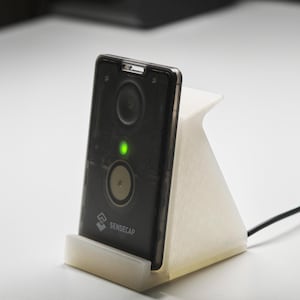 Peut inclure: Un appareil électronique noir avec deux composants circulaires et une lumière verte, posé dans un support blanc. L'appareil est relié à un câble noir et porte le logo "SENSECAP". Le support est sur une surface blanche.