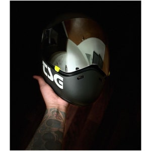 Pu&ograve; includere: Un casco da moto nero con una visiera trasparente e un logo "TSG" bianco sul lato. Il casco &egrave; tenuto in mano da una persona.