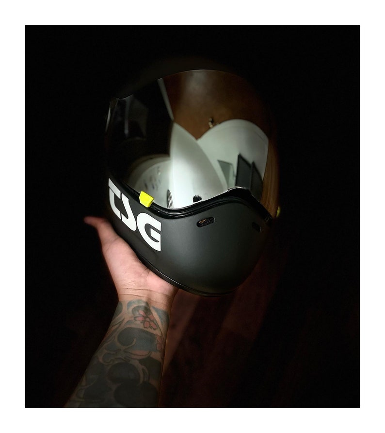 Pu&ograve; includere: Un casco da moto nero con una visiera trasparente e un logo "TSG" bianco sul lato. Il casco &egrave; tenuto in mano da una persona.