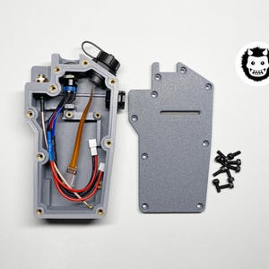 May include: A grey electronic device, partially disassembled, revealing internal wiring and a black cap. A separate grey panel, a pile of black screws, and a monster face sticker are also visible.