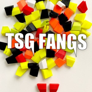 Pu&ograve; includere: Una collezione di tasti in plastica colorati in giallo, arancione, rosso, nero e bianco. I tasti sono disposti in un mucchio e presentano il testo "TSG FANGS".
