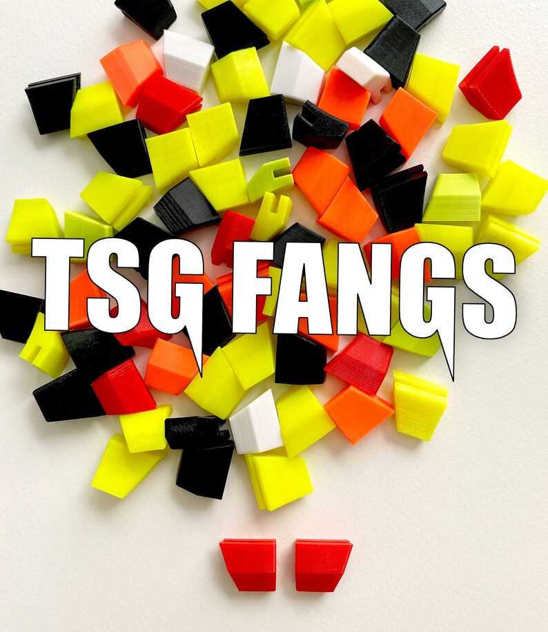 Pu&ograve; includere: Una collezione di tasti in plastica colorati in giallo, arancione, rosso, nero e bianco. I tasti sono disposti in un mucchio e presentano il testo "TSG FANGS".