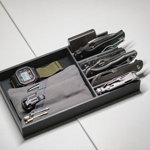 Könnte beinhalten: Ein schwarzes, rechteckiges Ablagefach mit einer Digitaluhr mit olivgrünem Armband, einem grauen Stoffbeutel, einem Metallfeuerzeug, drei Klappmessern, einem Multifunktionswerkzeug, einem Metallstift und zwei kleinen zylindrischen Metallteilen.