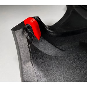 May include: Close-up of a black plastic object with a red plastic tab. The object has a textured surface and a sharp edge.