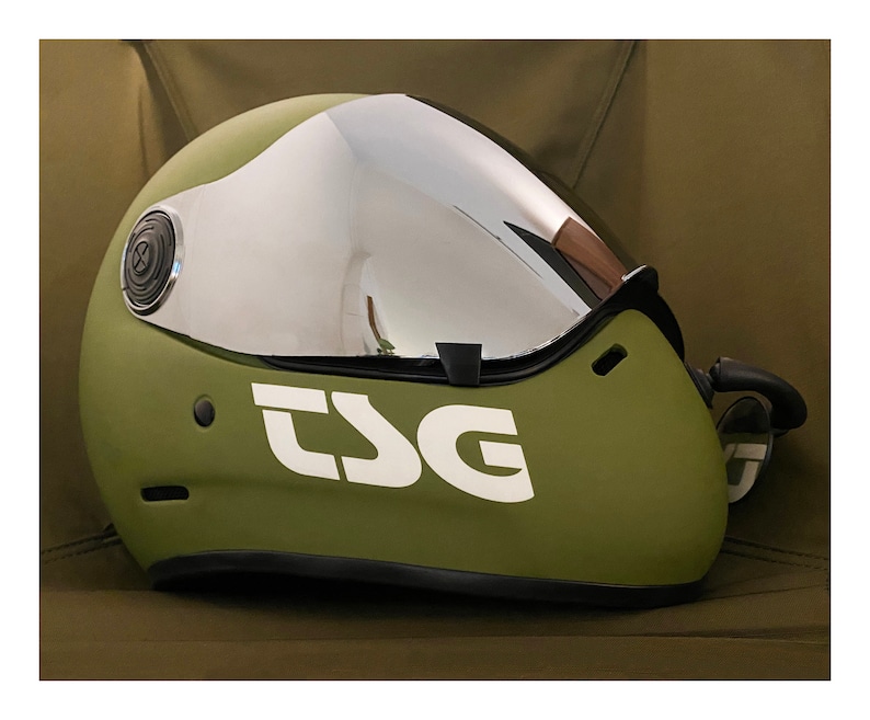 Pu&ograve; includere: Un casco verde con una visiera argentata e la scritta "TSG" in lettere bianche sul lato.