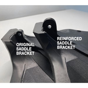 May include: Close-up of two black plastic saddle brackets. The bracket on the left is labeled "ORIGINAL SADDLE BRACKET" and the bracket on the right is labeled "REINFORCED SADDLE BRACKET".