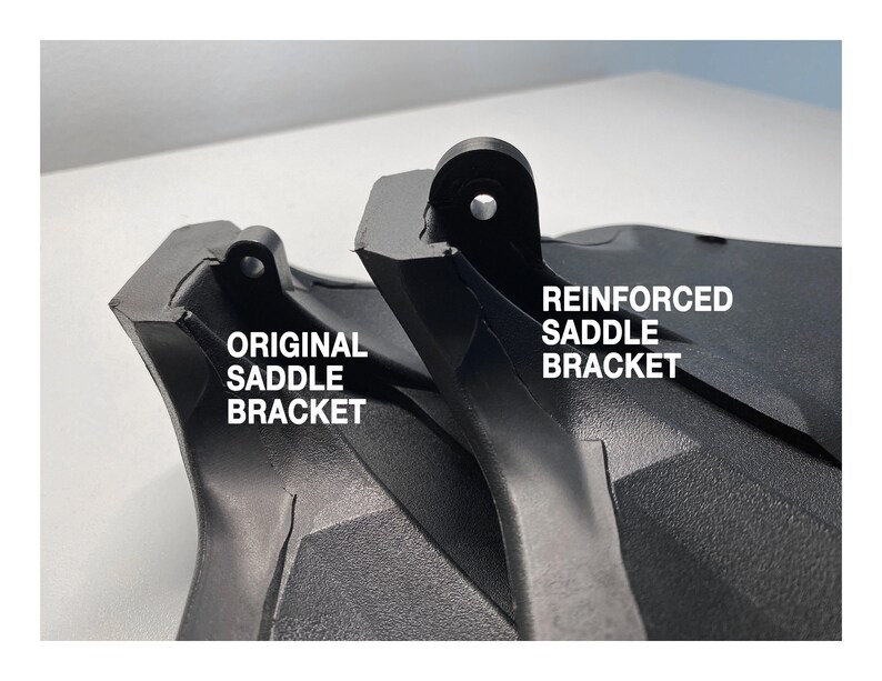 May include: Close-up of two black plastic saddle brackets. The bracket on the left is labeled "ORIGINAL SADDLE BRACKET" and the bracket on the right is labeled "REINFORCED SADDLE BRACKET".