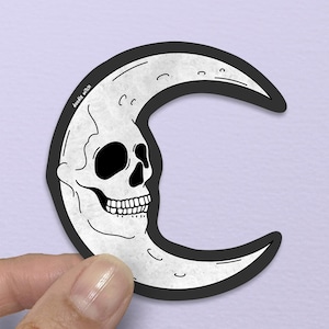 以下が含まれることがあります： 黒と白のステッカーで、ドクロの顔をした三日月が描かれています。ドクロには詳細な眼窩、鼻腔、完全な歯並びがあります。ステッカーは黒い縁取りがあり、紫色の背景に保持されています。