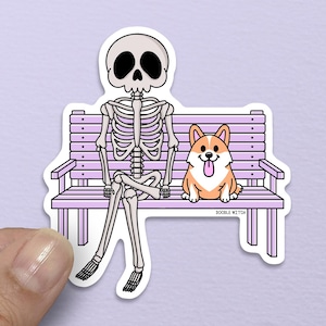 Puede incluir: Una pegatina con un esqueleto de dibujos animados y un perro corgi sentados en un banco de color lila claro. El esqueleto está sentado con las piernas cruzadas, y el corgi está a su lado con la lengua fuera. La pegatina tiene un borde blanco.