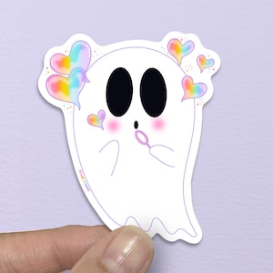May include: A white ghost sticker with black eyes and rosy cheeks. The ghost is blowing a bubble and has rainbow heart accents. The sticker is held by a hand against a light purple background.
