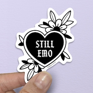 May include: A black and white sticker featuring a heart with the words "STILL EMO" in a gothic font. The heart is surrounded by floral and leaf designs. The sticker is held by a hand against a purple background.