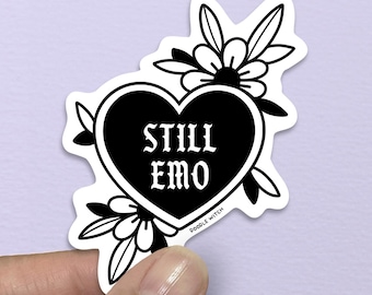 May include: A black and white sticker featuring a heart with the words "STILL EMO" in a gothic font. The heart is surrounded by floral and leaf designs. The sticker is held by a hand against a purple background.