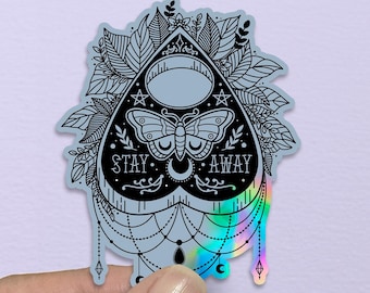 May include: A holographic sticker in the shape of a planchette, featuring a butterfly, crescent moons, and the words "STAY AWAY." The sticker has a black and silver design with iridescent accents and decorative foliage.