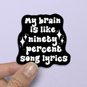 May include: A black sticker with white text that reads "My brain is like ninety percent song lyrics." The sticker has a wavy, irregular shape and is held by a hand against a light purple background.