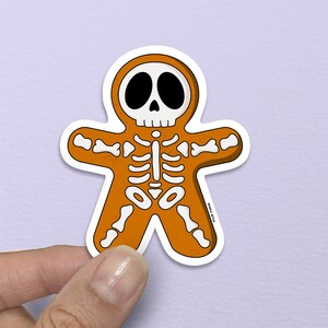 May include: A Halloween-themed sticker in the shape of a gingerbread man, featuring a skeleton design. The sticker is orange with white and black details, held against a light purple background.
