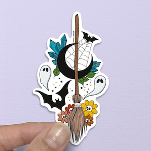 May include: A colorful sticker featuring a witch's broom, a crescent moon, a spider web, bats, ghosts, and flowers. The sticker has a white border and is held by a hand against a purple background. The text "DOODLE WITCH" is visible.