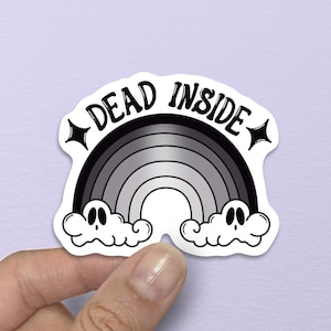 May include: A sticker with a black and white rainbow design and the words "DEAD INSIDE". The rainbow has a gradient effect, and the clouds have cartoon faces. The sticker is held by a hand.