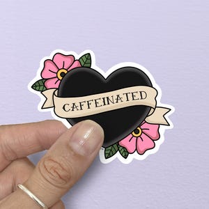 May include: A black heart-shaped sticker with the word "CAFFEINATED" on a banner. The sticker is decorated with pink flowers and green leaves. The sticker is held by a hand.
