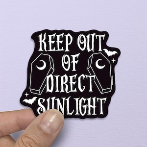 Pode incluir: Adesivo preto com o texto branco "KEEP OUT OF DIRECT SUNLIGHT", apresentando ilustrações de caixões e morcegos. O adesivo é segurado por uma mão contra um fundo roxo claro.