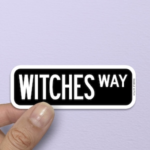 May include: A black and white sticker with the words "WITCHES WAY" in a bold, sans-serif font. The sticker has a white border and is held by a hand against a light purple background. The sticker is rectangular with rounded corners.