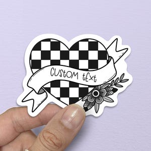 May include: A black and white sticker in the shape of a heart with a checkered pattern. A banner across the heart reads "CUSTOM TEXT". The sticker is decorated with a floral design.